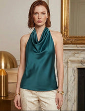 Woman wearing a teal halter top in an elegant room with a fireplace and gold lamp. Deep teal silk satin halter top with fluid drape and elegant halter neckline, luxury evening style