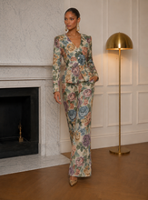 Woman in a floral Bella rose couture suit standing in a room with a fireplace and gold lamp.