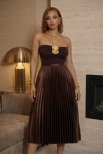 Woman in a brown strapless dress with a gold emblem standing in a room with a lamp and fireplace.  The Lumière Plissé Skirt is a high-waisted luxury pleated midi skirt designed with fluid movement and timeless elegance. Available in rich Espresso and refined Crème for elevated day-to-night styling.