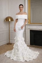 Woman in a white off-shoulder gown with floral details in an elegant room.

The La Roseraie Gown is an exquisite expression of modern couture and timeless femininity. Designed to sculpt and flatter, this off-the-shoulder mermaid silhouette elegantly frames the neckline while contouring the waist and hips for a refined, elongated look.

Crafted with a structured bodice and smooth, body-skimming fabrication, the gown flows effortlessly into a dramatic floor-length train adorned with hand-placed three-dimensio